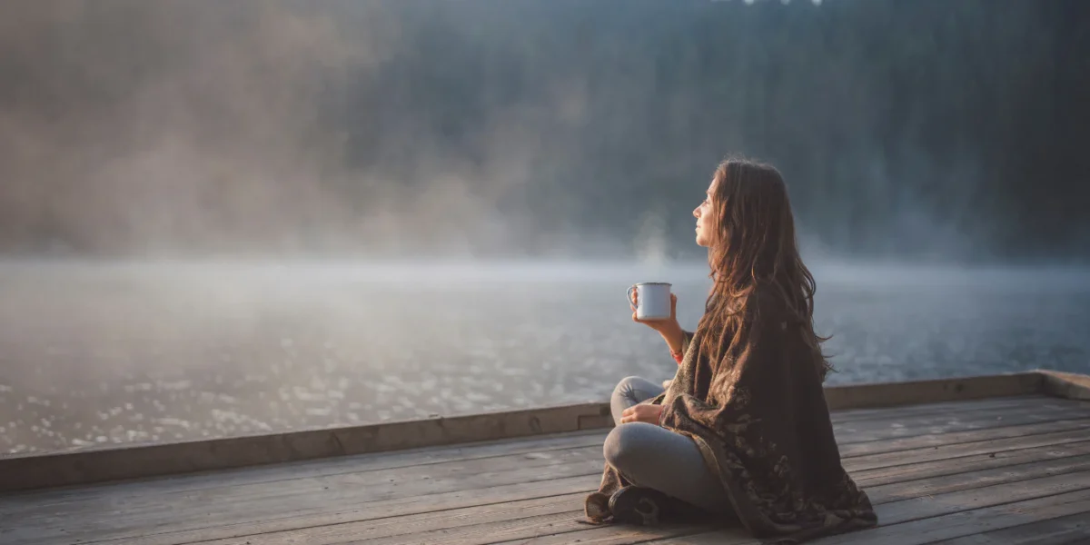 Woman sitting with a cup on a dock beside a misty lake