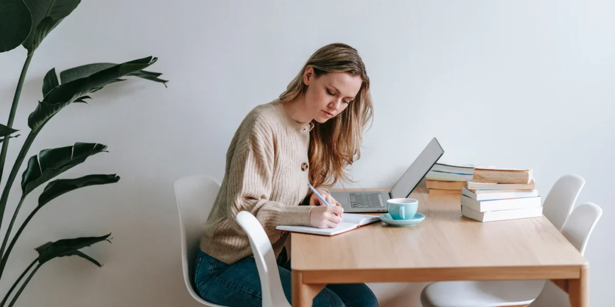 Person writing at a table beside a laptop and books